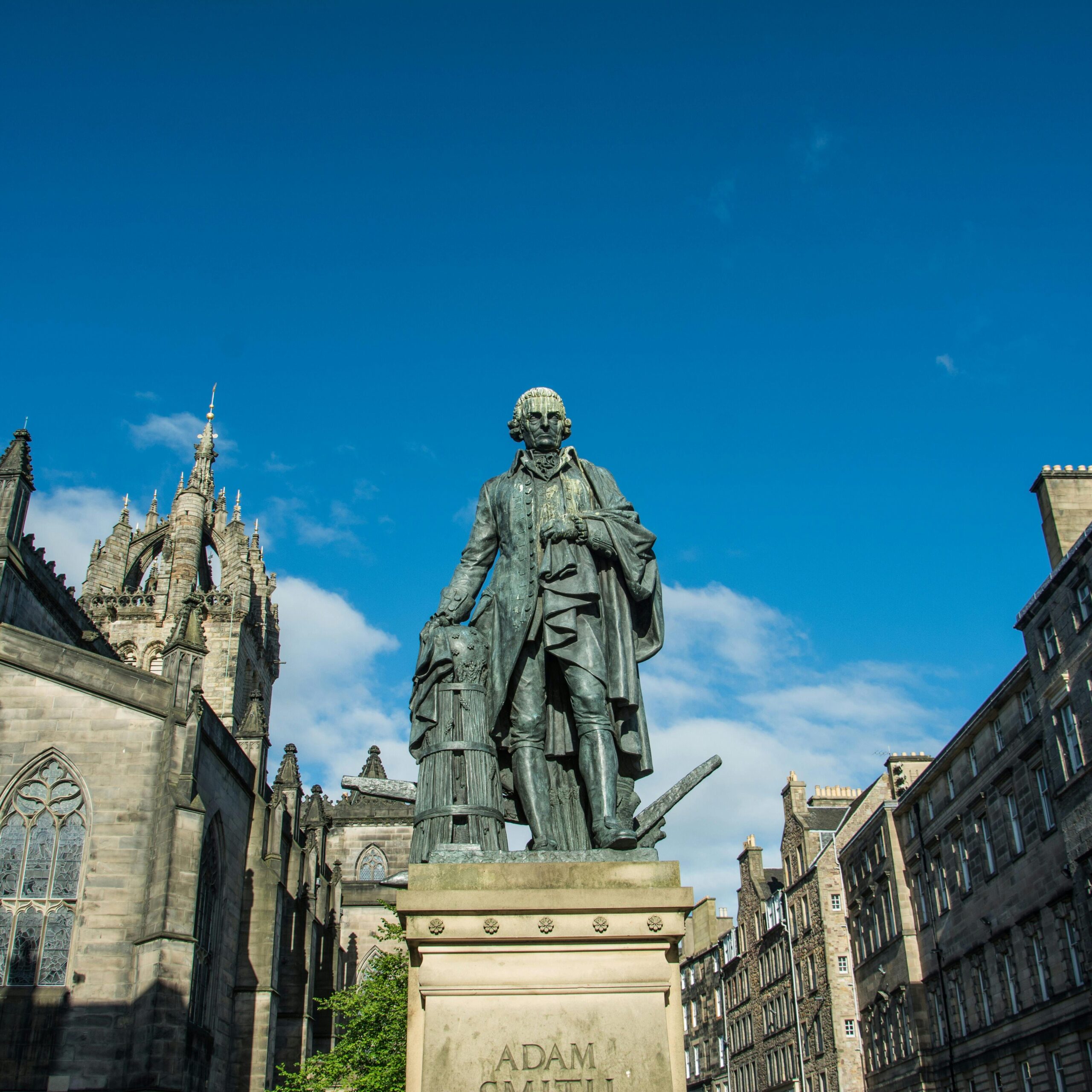 Statue of Adam Smith with historic buildings in Edinburgh, Scotland, under a blue sky.