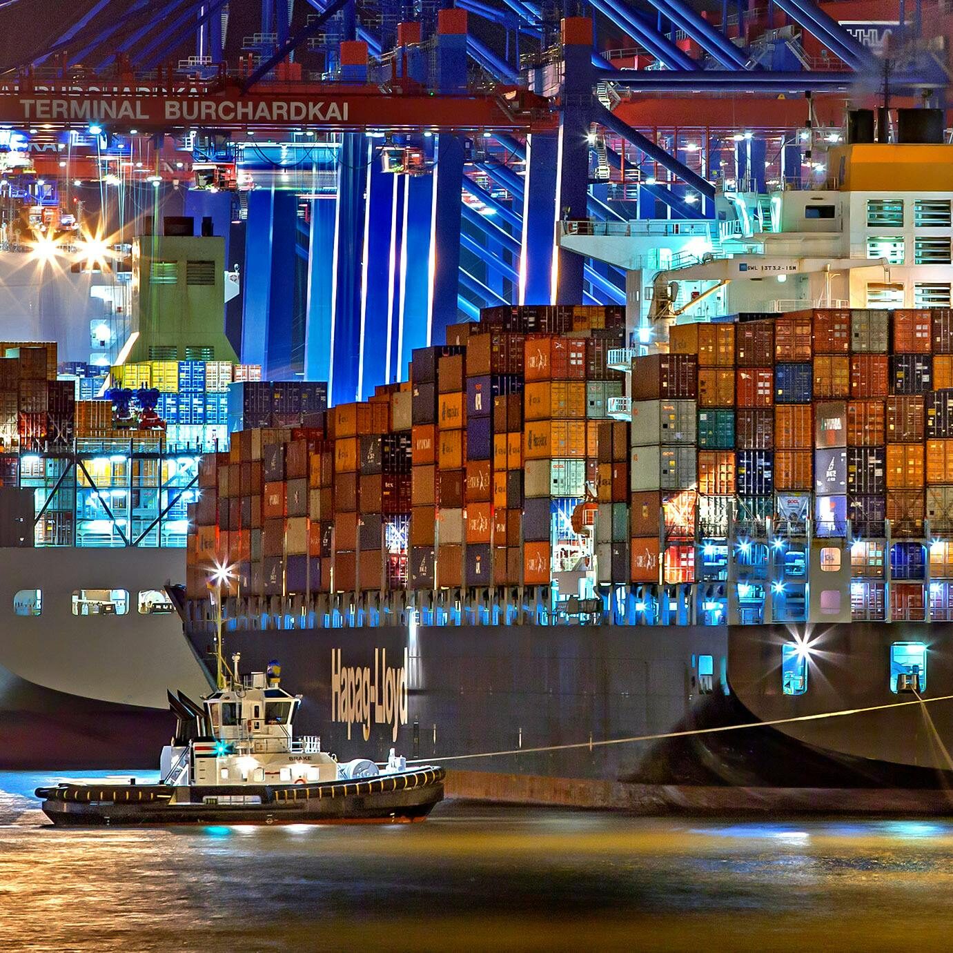 A brightly lit cargo ship at Hamburg harbor with stacked containers and a tugboat.
